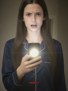 A woman in dark pyjamas, lit from below by her phone, staring past the camera in a dim hallway.